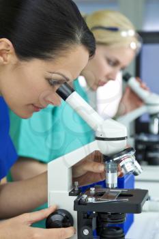 A female medical or scientific researcher or doctor using her microscope in a laboratory with her blond colleague out of focus behind her.