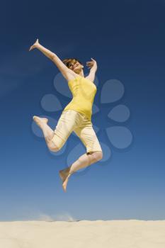 Photo of joyful female in casual clothes jumping over sand with her arms raised