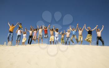 Crowd of friends jumping on sandy beach with their arms raised against blue sky