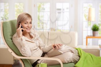 Cheerful woman speaking on cellphone sitting in living room armchair.