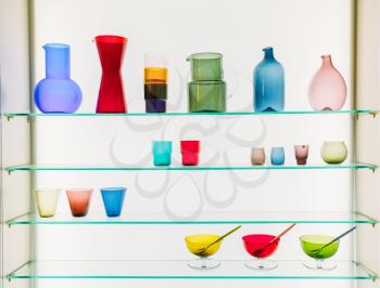Assorted Different Sizes And Shapes Of Colorful Glassware On Shelves. Vases, Pitchers, Jugs, Glasses On White Background.