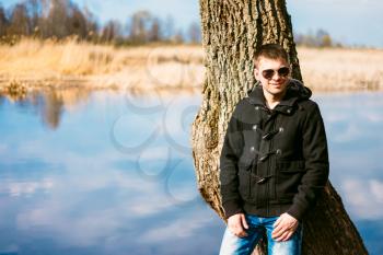Young Handsome Man Leaned Against A Tree By River In Autumn Day, Smiling In Camera. Casual Style - Jeans, Jacket, Sunglasses
