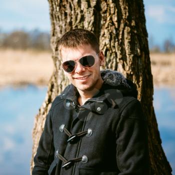 Young Handsome Man Leaned Against A Tree By River In Autumn Day, Smiling In Camera. Casual Style - Jacket, Sunglasses