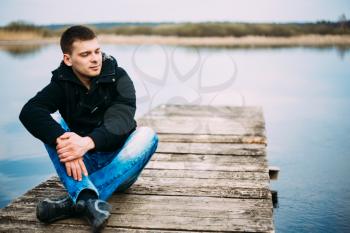 Young Handsome Man Sitting On Wooden Pier In Autumn Day, Relaxing,  Thinking, Listening. Casual Style - Jeans, Jacket