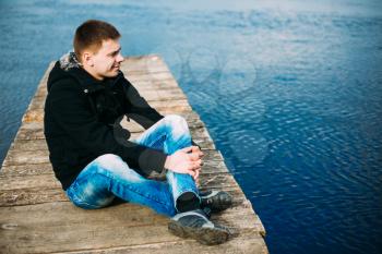 Young Handsome Man Sitting On Wooden Pier In Autumn Day, Relaxing,  Thinking, Listening. Casual Style - Jeans, Jacket