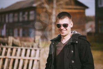 Young Handsome Man Staying Near Old Wooden House In Autumn Day, Relaxing,  Thinking, Smiling. Casual Style - Jacket, Sunglasses.