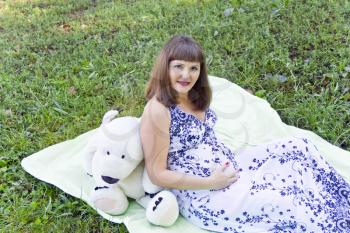 Pregnant woman with big white toy bear sitting on green grass