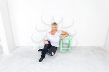 Blond woman in white and black jeans sitting on the stool inside empty room