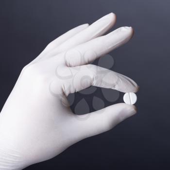 Hand in white latex glove holding white pill on dark background