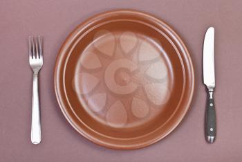 top view of empty ceramic brown plate with fork and knife set on brown background