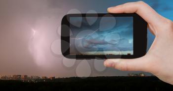 travel concept - tourist takes picture of lightning bolt in storm clouds over city in summer evening on smartphone, Moscow, Russia