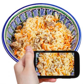 photographing food concept - tourist takes picture of traditional asian pilau with meat in ceramic bowl on smartphone, Uzbekistan