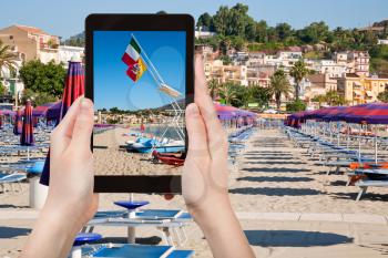 travel concept - tourist takes picture of flags Italy and Sicily at urban sand beach in resort Giardini Naxos, Sicily, Italy on tablet pc