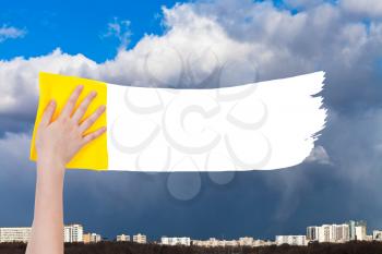 weather concept - hand deletes rainy clouds over city by yellow rag from image and white empty copy space are appearing