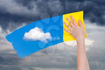 weather concept - hand deletes rain clouds by yellow cloth from image and blue sky with white cloud is appearing