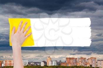 weather concept - hand deletes rainy clouds over urban houses by yellow rag from image and white empty copy space are appearing