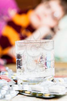 glass with dissolved medicament in water and pile of pills on table close up and sick girl with scarf around her neck on sofa in living room on background