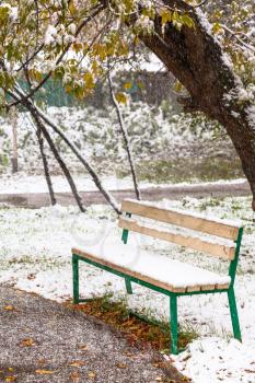 first snowfall and empty bench in city park in autumn day