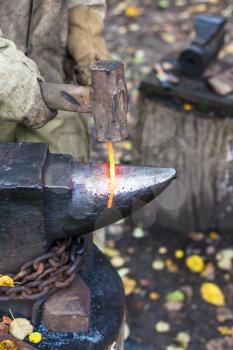 Blacksmith forges red hot glowing iron rod on anvil in outdoor rural smithy