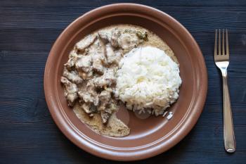 Russian cuisine dish - served portion of Beef Stroganoff (Beef Stroganov, Befstroganov) pieces of stewed meat in sour cream with boiled rice on brown plate on dark table