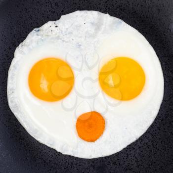 fried eggs and slice of carrot on black plate close up. Fried eggs like face with open mouth