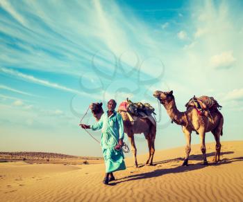Vintage retro hipster style travel image of Rajasthan travel background - Indian cameleer (camel driver) with camels in dunes of Thar desert. Jaisalmer, Rajasthan, India
