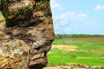 Old rock with lichen looks like the human face against of meadow and blue sky.