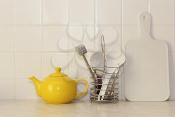 Yellow ceramic teapot and kitchen utensil on white kitchen table.
