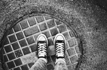 Teenager feet in sneakers. Gumshoes standing an urban manhole cover. Closeup black and white photo with selective focus, old style