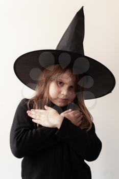 Angry little blond European girl in black witch costume over white wall, close-up studio portrait