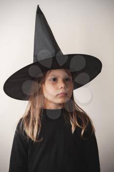 Serious little blond European girl in black witch costume over white wall, close-up studio portrait