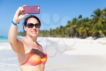 Smiling young Caucasian woman in bright swimsuit and sunglasses takes selfie photo on the beach of Saona island, Dominican Republic