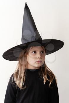 Little blond European girl in black witch costume stands near white wall, close-up studio portrait