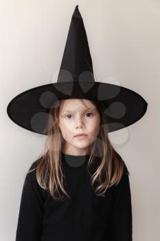 Little blond European girl in black witch costume posing over white wall, close-up studio portrait