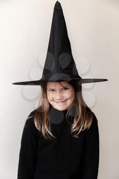 Smiling little blond European girl in black witch costume over white wall, close-up studio portrait