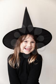 Little blond girl in black witch costume smiles over gray wall, close-up studio portrait