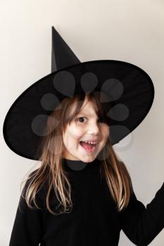 Smiling little blond European girl in black witch costume over white wall, close-up portrait