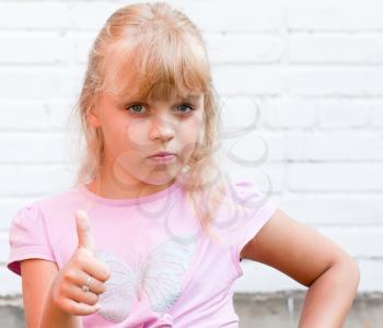 Portrait of a little blond beautiful Russian girl with thumbs up above white brick wall background