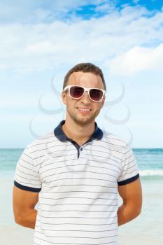 Outdoor portrait of young strong smiling Caucasian man standing with white sunglasses on summer sea coast