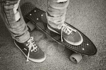 Young skateboarder in gumshoes and jeans standing on his skate. Close-up fragment of skateboard and feet, monochrome retro stylized photo