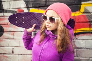 Blond teenage girl holds skateboard near by urban wall with graffiti