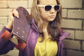 Blond teenage girl in sunglasses holds skateboard near gray urban brick wall, vintage warm tonal correction, old style photo filter effect