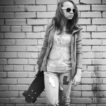 Blond teenage girl in jeans and sunglasses holds skateboard over urban brick wall background, monochrome photo