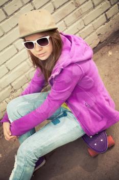 Blond teenage girl in cap and sunglasses sits on her skateboard near urban brick wall, vertical photo with warm retro tonal correction effect, instagram old style filter