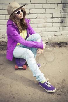 Blond teenage girl in jeans and sunglasses sits on her skateboard near urban brick wall, vertical photo with warm retro tonal correction effect, instagram old style filter