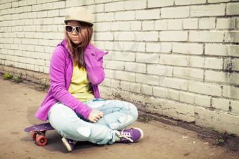 Blond teenage girl in jeans and sunglasses sits on her skateboard near urban brick wall, photo with warm retro tonal correction effect, instagram old style filter