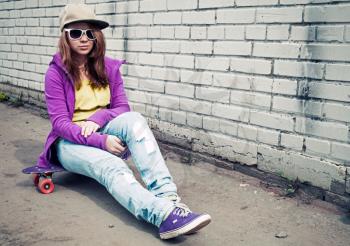 Blond teenage girl in jeans and sunglasses sits on her skateboard near urban brick wall, photo with cold retro tonal correction effect, instagram old style filter