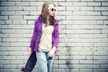 Blond teenage girl in jeans and sunglasses holds skateboard near gray urban brick wall, cold vintage tonal correction, old style instagram photo filter effect