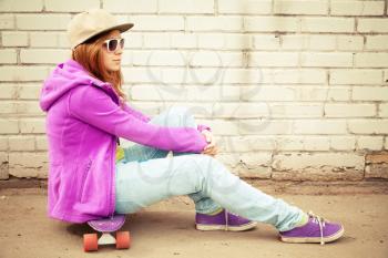 Blond teenage girl in jeans, cap and sunglasses sits on her skateboard near urban brick wall, photo with warm retro tonal correction effect, instagram old style filter