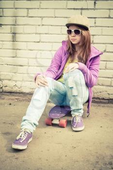 Beautiful teenage girl in cap and sunglasses sits on a skateboard near urban brick wall, vertical photo with warm retro tonal correction effect, instagram old style filter